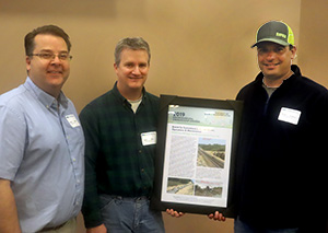 Picture of three men, with the middle man holding an award plaque.